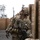 A heavily equipped soldier in MultiCam/OCP camouflage uniform is positioned within a wooden training lane or mock urban environment, looking upward while holding an assault rifle. The soldier is wearing full combat gear including a helmet with camouflage netting and mounted devices, tactical vest with radio equipment, and protective eyewear. The setting appears to be a military training facility with constructed wooden wall corridors.