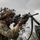 A U.S. Army soldier in full combat gear is shown aiming a rifle in a field environment. The soldier is wearing MultiCam (OCP) camouflage uniform with a ghillie-style helmet cover, face paint, and tactical accessories. The image appears to be taken during a training exercise or field operation.
