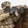 A fully equipped soldier in MultiCam/OCP camouflage uniform is positioned behind a wooden barrier in a tactical shooting stance. The soldier is aiming an M4/M16-style carbine equipped with optics and accessories over the barrier. The setting appears to be a training range or exercise environment based on the constructed wooden obstacle.