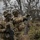 Two U.S. Army soldiers in full combat gear are conducting field operations in a wooded, rocky terrain. One soldier is pointing in a direction, apparently giving or receiving tactical guidance, while the other holds an assault rifle at the ready. Both wear MultiCam uniforms, tactical helmets with foliage camouflage, face paint, and full kit including body armor and accessories.