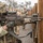 A U.S. Army soldier identified by name tape as 'SZOSTECKI' is seen aiming an M249 Squad Automatic Weapon (SAW) around a wooden barrier during what appears to be a training exercise. The soldier is wearing full OCP (Operational Camouflage Pattern) kit including a camouflage helmet cover, tactical vest, and gloves. Additional soldiers in similar gear are visible in the background along a wooden fence line.