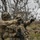 Two soldiers in full combat gear and camouflage uniforms are engaged in a tactical coordination exercise in a rugged outdoor environment. One soldier is pointing in a specific direction while the other observes, suggesting target designation or navigation coordination. The setting appears to be a training exercise based on the terrain and equipment configuration.