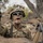 A soldier in full combat gear is shouting and pointing, appearing to issue commands or direct movement during what appears to be a field exercise or operation. The soldier is wearing camouflage uniform, tactical helmet with accessories, face paint, and dark ballistic eyewear. A second soldier is partially visible to the right holding a rifle.