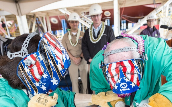 Two individuals wearing patriotic American flag-themed welding helmets and green protective jackets are performing a ceremonial welding operation on a metal plate, likely a keel-laying or ship-naming ceremony. Several observers wearing hard hats and Hawaiian leis stand in the background, suggesting a formal maritime shipbuilding event. The active welding spark is visible at the point of contact with the metal surface.