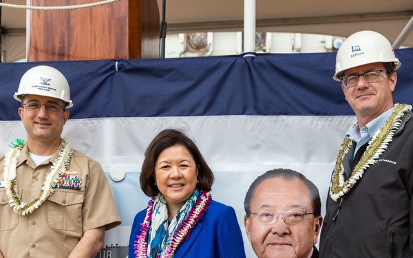 Three individuals pose in front of a banner at what appears to be a naval ship keel-laying ceremony hosted by General Dynamics Bath Iron Works. A military officer in uniform and a civilian in a black jacket wearing hard hats flank a woman in a blue suit, all wearing Hawaiian leis. A large portrait of an Asian man is displayed on the banner behind them.