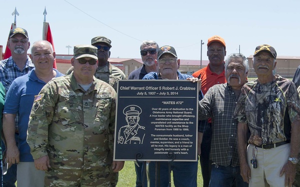 A group of approximately 12-14 individuals, including active-duty military personnel in camouflage uniforms and civilians, pose around a memorial plaque dedicated to Chief Warrant Officer 5 Robert J. Crabtree (July 5, 1937 – July 3, 2014). The plaque, nicknamed 'MATES #72,' honors his over 40 years of dedication to the Oklahoma Army National Guard. The ceremony appears to be a formal dedication event held outdoors on a military installation.