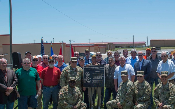 A group of approximately 25-30 people, including both military personnel in camouflage uniforms and civilians, are gathered outdoors at what appears to be a military installation for a memorial or dedication ceremony. They are posed around a mounted commemorative plaque dedicated to Chief Warrant Officer 5 Robert J. Crabtree. Military flags and base buildings are visible in the background.