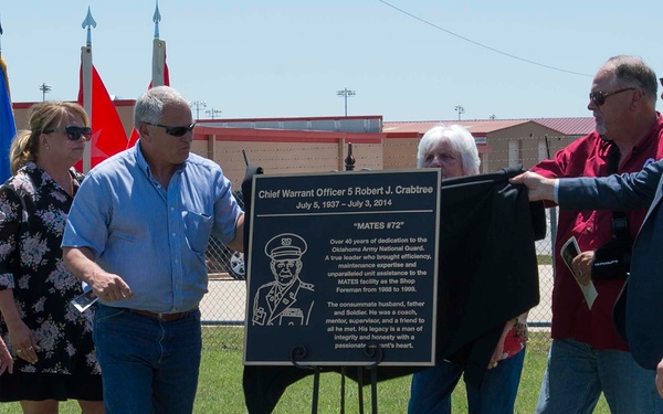 A group of six people are gathered outdoors at what appears to be a military installation for a memorial plaque unveiling ceremony honoring Chief Warrant Officer 5 Robert J. Crabtree (July 5, 1937 – July 3, 2014). The black commemorative plaque, mounted on an easel, bears the nickname 'MATES #72' and references his over 40 years of dedication to the Oklahoma Army National Guard. Two men on the right side are in the process of removing a dark cloth covering from the plaque.