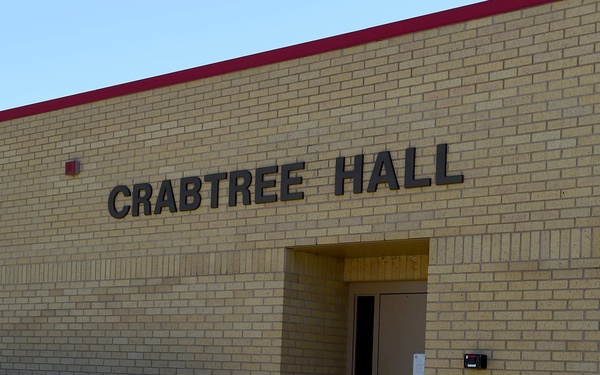 The image shows the exterior facade of a building named 'Crabtree Hall,' constructed with light tan/yellow brick and featuring a dark red accent band along the roofline. A single entrance door is visible at the center, flanked by trimmed green shrubs at ground level. The building appears to be an institutional or academic structure, likely part of a university or college campus.