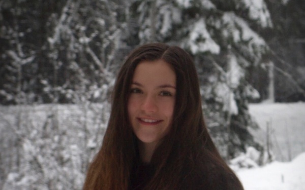 A young woman with long dark brown hair is posing outdoors in a snowy winter setting. She is smiling at the camera while wearing a dark jacket and light blue jeans. Snow-covered evergreen trees are visible in the background.