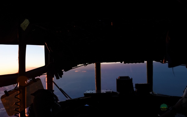 The image shows the interior of a military aircraft cockpit with two crew members silhouetted against a dramatic sunset visible through the multi-pane windscreen. The aircraft appears to be a large military transport or patrol aircraft, likely a C-130 Hercules based on the distinctive cockpit window configuration. The scene is captured at high altitude with cloud layers visible below.