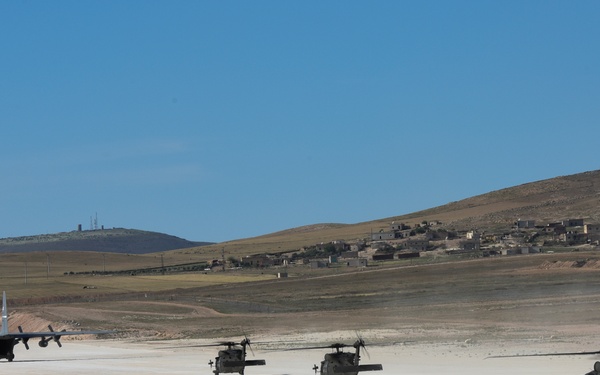 A military airfield scene showing three UH-60 Black Hawk helicopters lined up on a dirt/gravel runway alongside a fixed-wing transport aircraft, likely a C-130 Hercules. The setting appears to be an arid, semi-desert environment with rolling hills, sparse vegetation, and a small settlement visible in the background. Two personnel are visible on foot near the fixed-wing aircraft.