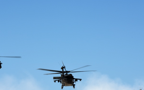 Three military helicopters are flying in formation against a clear blue sky, partially obscured by white smoke or dust clouds in the lower portion of the image. The helicopters appear to be utility/transport type aircraft consistent with UH-60 Black Hawk design. The scene suggests a military operation, exercise, or demonstration involving rotary-wing aircraft.
