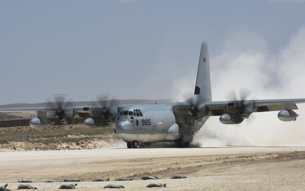 A military C-130 Hercules transport aircraft marked '985' is landing or taking off on an unpaved desert airstrip, kicking up significant dust clouds. The aircraft appears to be a gray military variant operating in an arid, desert environment. Sand bags or supply bundles are visible along the edge of the runway.