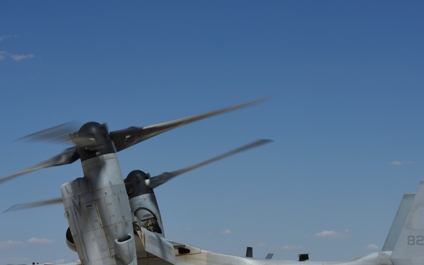 A U.S. Marines MV-22 Osprey tiltrotor aircraft (bureau number 8296, tail code YT 06) is on the ground with rotors spinning at what appears to be a forward operating base or austere airstrip in an arid environment. A group of approximately 8-10 armed military personnel in combat gear are standing near the aircraft, likely preparing to board or having just disembarked. The terrain and landscape suggest a desert or semi-arid operational environment.