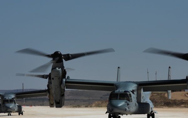 Three military tiltrotor aircraft, identifiable as V-22 Osprey variants, are positioned on a sandy/arid airstrip with rotors spinning. The aircraft are in a ground taxi or pre-takeoff configuration, with nacelles tilted upward in helicopter mode. The setting appears to be a desert or arid region based on the terrain and hazy sky.
