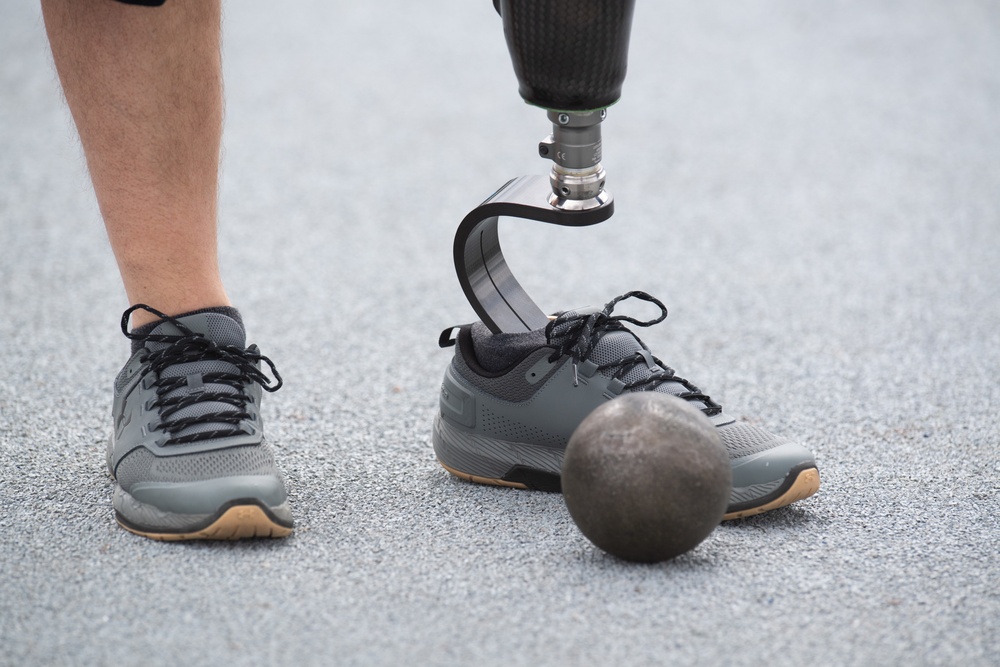 The image shows the lower legs of a person, one of which is a prosthetic limb made of carbon fiber with a curved blade design and metallic connector joint. Both legs are wearing matching gray athletic sneakers with black laces and gum soles. A dark, worn spherical ball — likely a shot put or medicine ball — rests on the ground near the prosthetic foot.