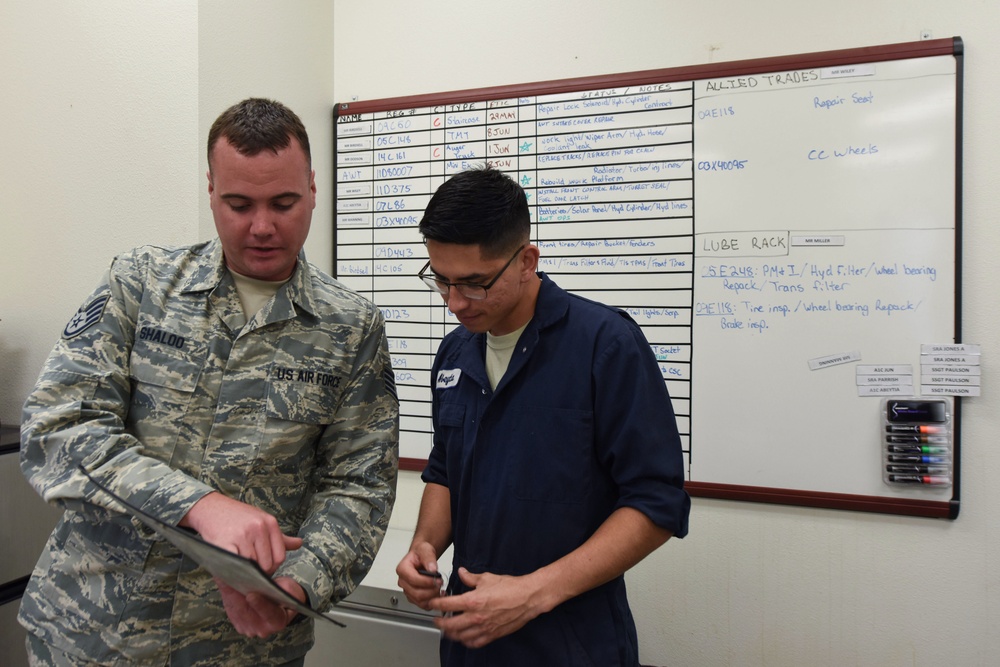 Two military personnel are reviewing documents together in what appears to be a maintenance or operations office. One individual is wearing US Air Force ABU (Airman Battle Uniform) with the name tape 'SHALDO' visible, while the other is wearing a blue work uniform. A whiteboard behind them displays maintenance scheduling information including sections labeled 'Allied Trades' and 'Lube Rack'.