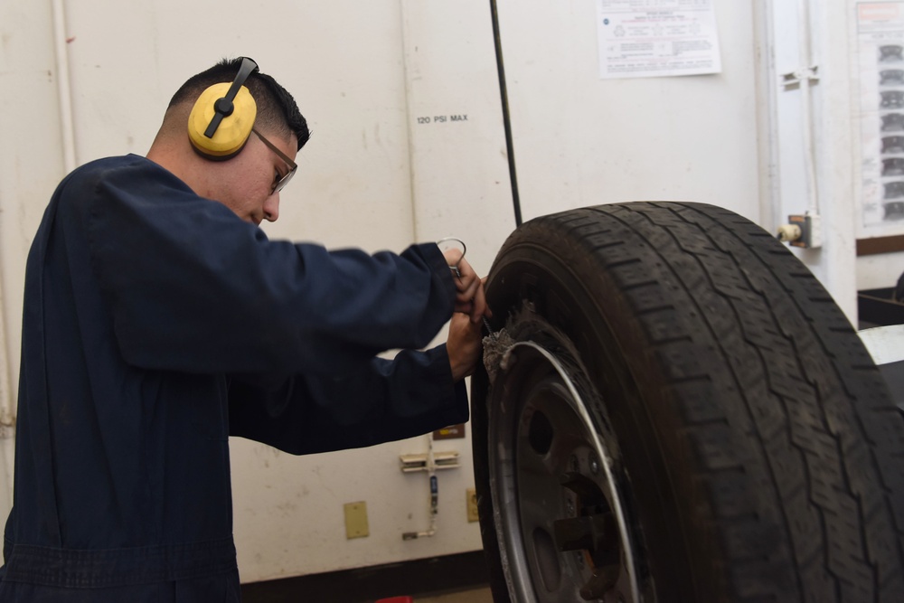 A mechanic wearing yellow ear protection and safety glasses is working on a large tire in what appears to be an automotive or fleet maintenance shop. He is dressed in a dark blue work uniform and is closely inspecting or servicing the wheel/tire assembly. A label on the wall behind him reads '120 PSI MAX', indicating a pressurized air system nearby.