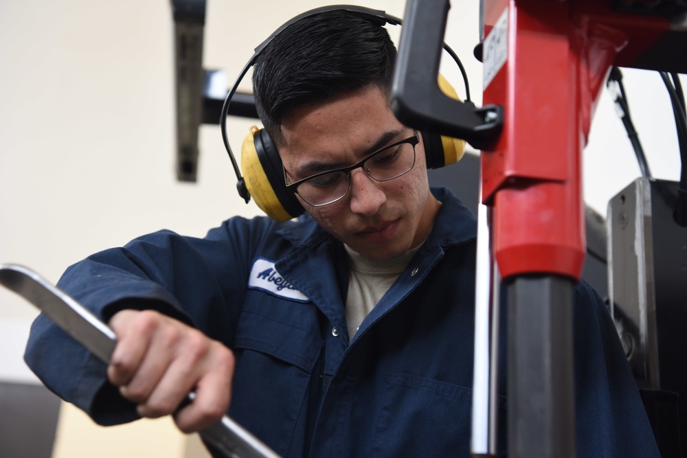 A young male technician or mechanic is focused on operating or adjusting industrial machinery in what appears to be a workshop or manufacturing environment. He is wearing appropriate personal protective equipment including yellow ear defenders and glasses. His navy blue work uniform bears a partially visible logo or name patch.