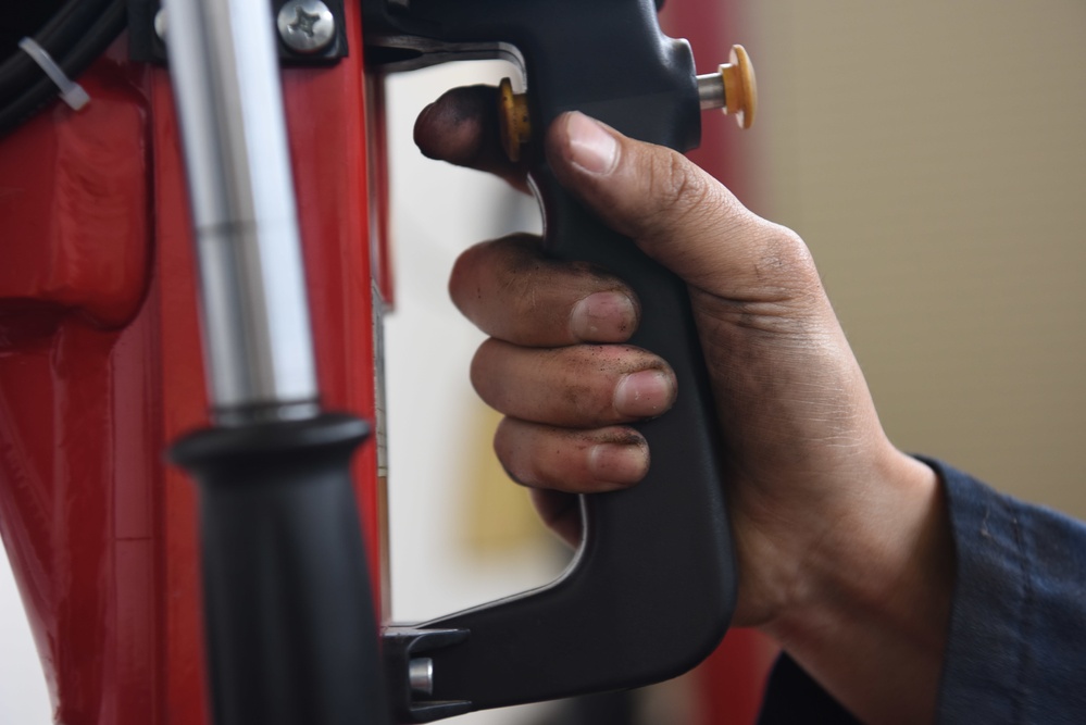 A close-up photograph of a person's hand gripping what appears to be a motorcycle mirror assembly on a red motorcycle. The hand shows signs of mechanical work, with visible dirt/grease on the fingers and knuckles. The image captures a maintenance or assembly task being performed on the vehicle.