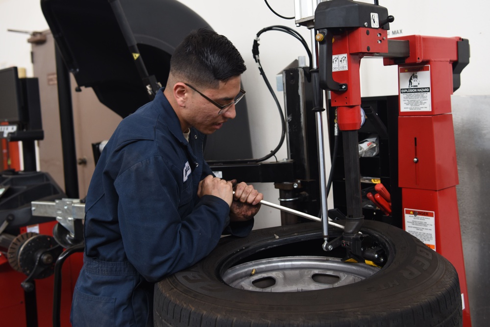 A mechanic in blue coveralls is operating a red tire changing machine in an automotive service bay. He is using a tire iron/bar to mount or dismount a tire from a wheel rim on the machine. The workspace appears to be a professional garage with additional equipment visible in the background.