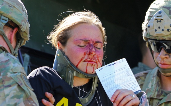 A military medical training exercise showing two soldiers in OCP (Operational Camouflage Pattern) uniforms attending to a simulated casualty. The 'casualty' is a woman wearing an Army PT shirt with simulated facial injuries (moulage makeup) and a cervical collar, holding what appears to be a casualty card. This appears to be a TCCC (Tactical Combat Casualty Care) or mass casualty training scenario.