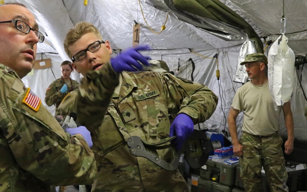 U.S. Army medical personnel are conducting what appears to be a field medical training exercise inside a military tent. Two soldiers in OCP camouflage uniforms with medical gloves are attending to a simulated casualty lying on a stretcher, who has theatrical makeup simulating facial injuries. Additional soldiers observe the exercise in the background near military medical supply cases.