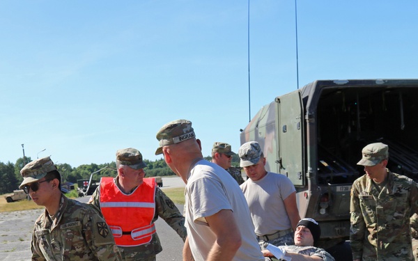 A group of U.S. Army soldiers in OCP (Operational Camouflage Pattern) uniforms are carrying a casualty on a litter/stretcher across a paved surface near a military vehicle. One individual is wearing a high-visibility orange safety vest, suggesting an observer or safety officer role. The scene appears to be a medical evacuation training exercise based on the controlled environment and observer presence.