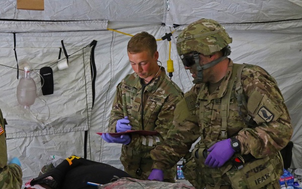 U.S. Army soldiers are conducting a medical training exercise inside a military field hospital tent. Three soldiers in OCP (Operational Camouflage Pattern) uniforms are treating a simulated casualty on a litter, with realistic moulage (simulated wounds) including a mock traumatic amputation. The scenario appears to be a combat casualty care training drill, with personnel wearing medical gloves and using documentation tools.