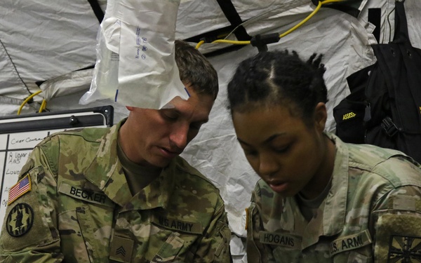 Two U.S. Army soldiers in OCP (Operational Camouflage Pattern) uniforms are providing medical care to a patient lying on a stretcher inside a military medical tent. The soldier on the left is identified by name tape as 'BECKER' and holds the rank of Sergeant, while the soldier on the right is identified as 'HOGANS' and is wearing blue medical gloves. This appears to be a field medical training exercise or operational medical treatment scenario.