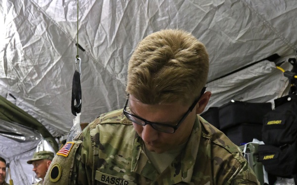 A U.S. Army soldier named BASSAR is providing medical treatment to a patient lying on a field stretcher inside a military medical tent. The soldier is wearing blue nitrile gloves and applying bandaging or wound care to the patient's head/face area. The setting appears to be a forward medical station or field hospital, likely during a training exercise or operational deployment.