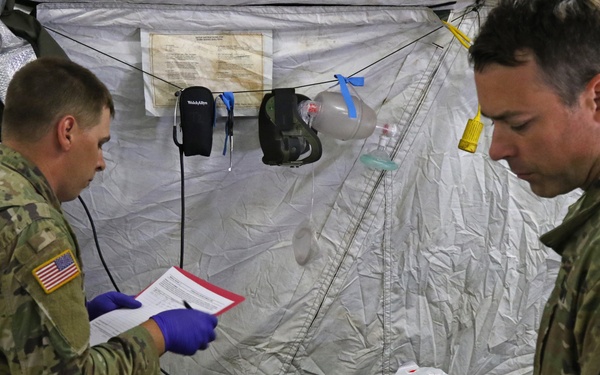Two U.S. Army soldiers in OCP (Operational Camouflage Pattern) uniforms are providing medical care to a patient lying on a stretcher inside a military field medical tent. One soldier is documenting information on a form while wearing purple nitrile gloves, and the other is operating a patient monitor/defibrillator device. The setting appears to be a field medical training exercise or operational medical treatment facility.