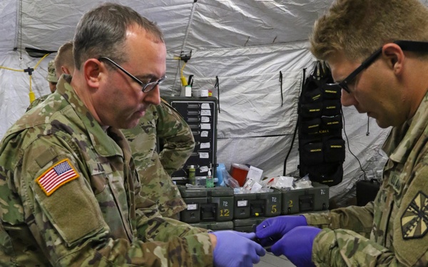 Two U.S. Army soldiers in MultiCam/OCP uniforms wearing purple nitrile gloves are providing medical treatment to a casualty inside a military field medical tent. The patient, wearing an Army PT shirt, is lying on a litter with head bandaging and an IV line in place. This appears to be a medical training exercise or field treatment scenario.