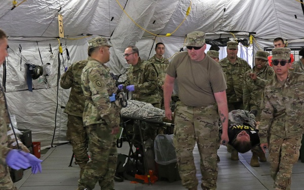 Military personnel in U.S. Army combat uniforms (OCP/MultiCam) are conducting what appears to be a medical training exercise inside a large military field hospital tent. Soldiers are transporting a simulated casualty on a litter/stretcher while others observe or assist. The setting includes medical equipment, IV stands, and supply containers consistent with a forward surgical or combat support hospital environment.