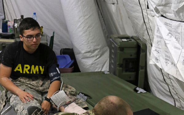 A U.S. Army medical scenario is taking place inside a military field tent. A soldier in OCP (Operational Camouflage Pattern) uniform wearing blue medical gloves is operating a medical monitoring device, while a second soldier wearing an 'ARMY' PT shirt and ACU pants sits on a field cot with a blood pressure cuff on his arm. The setting appears to be a field medical station or training exercise.