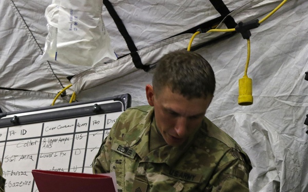 A U.S. Army soldier identified by nametape as 'BECKER' with the rank of Sergeant is providing medical treatment inside a military field tent or aid station. He is wearing blue medical gloves and attending to a patient's leg while referencing documents. A whiteboard tracking patient information is visible in the background.