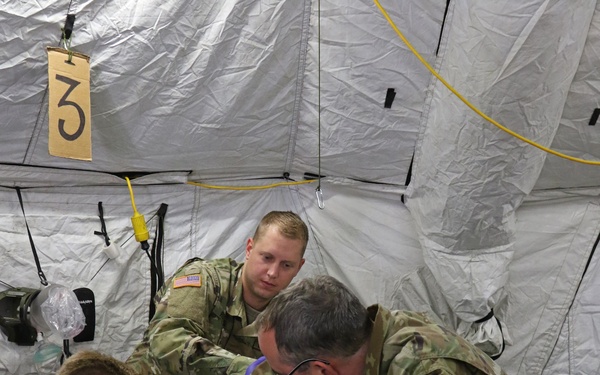 Three U.S. Army soldiers in MultiCam/OCP uniforms are performing a medical treatment procedure on a patient lying on a military field litter inside a military medical tent. Two soldiers wearing purple nitrile gloves are actively working on the patient while a third observes. The setting appears to be a field medical training exercise or operational medical station, indicated by the numbered station marker ('3') and modular tent structure.