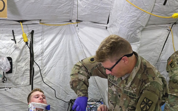 A U.S. Army medic identified by the name tape 'BASSAR' is providing medical treatment to a casualty lying on a military litter inside a field medical tent. The patient, wearing Army Combat Uniform (ACU pattern), has a cervical collar applied and appears to have simulated injuries including what appears to be a tourniquet on the leg. This appears to be a medical training exercise based on the controlled environment and visible simulation indicators.