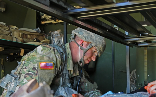 A U.S. Army soldier wearing MultiCam (OCP) uniform with a stethoscope is providing medical treatment to a casualty inside what appears to be a military medical vehicle or ambulance. The patient is lying on a litter and appears to have simulated wounds, suggesting this is likely a combat medical training exercise. The medic is actively assessing the patient using a stethoscope and appears to be monitoring vital signs.