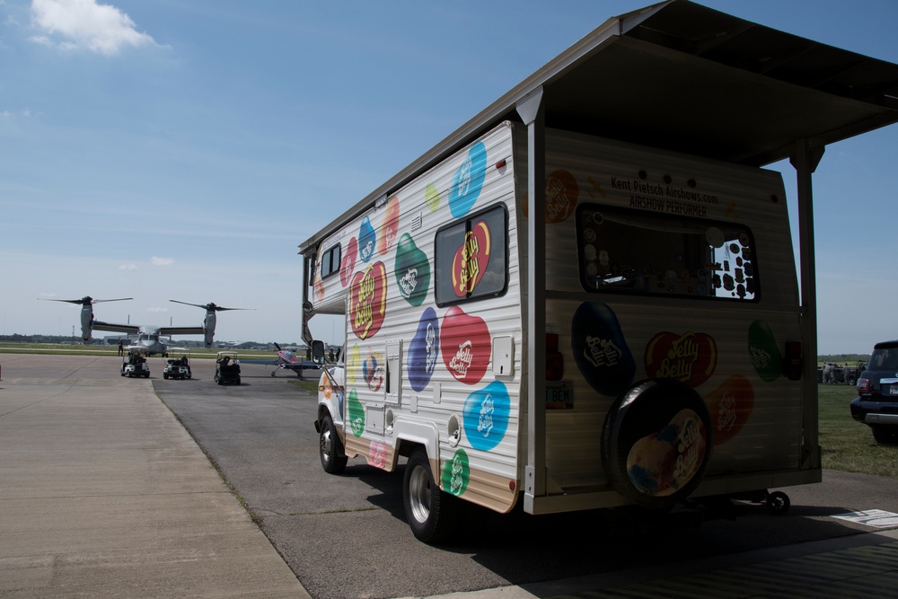 A Jelly Belly branded promotional vehicle is parked on an airfield tarmac at what appears to be an airshow. The vehicle is decorated with colorful Jelly Belly bean logos and branding, and has an open service window with an awning. In the background, a V-22 Osprey tiltrotor military aircraft is visible on the tarmac along with golf carts and other vehicles.
