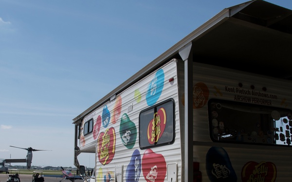 A Jelly Belly branded promotional vehicle is parked on an airfield tarmac at what appears to be an airshow. The vehicle is decorated with colorful Jelly Belly bean logos and branding, and has an open service window with an awning. In the background, a V-22 Osprey tiltrotor military aircraft is visible on the tarmac along with golf carts and other vehicles.