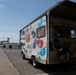 A Jelly Belly branded promotional vehicle is parked on an airfield tarmac at what appears to be an airshow. The vehicle is decorated with colorful Jelly Belly bean logos and branding, and has an open service window with an awning. In the background, a V-22 Osprey tiltrotor military aircraft is visible on the tarmac along with golf carts and other vehicles.