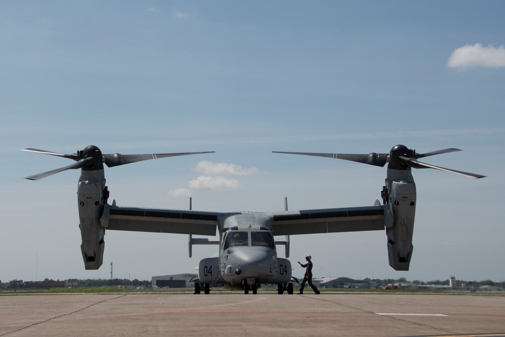 A Bell Boeing V-22 Osprey tiltrotor military aircraft is parked on a tarmac, viewed from a direct frontal perspective. The aircraft displays the number '04' on its nose and appears to be in a pre-flight or post-flight ground operation. At least one ground crew member is visible near the nose of the aircraft performing marshaling or inspection duties.
