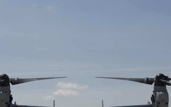 A Bell Boeing V-22 Osprey tiltrotor military aircraft is parked on a tarmac, viewed from a direct frontal perspective. The aircraft displays the number '04' on its nose and appears to be in a pre-flight or post-flight ground operation. At least one ground crew member is visible near the nose of the aircraft performing marshaling or inspection duties.