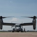 A Bell Boeing V-22 Osprey tiltrotor military aircraft is parked on a tarmac, viewed from a direct frontal perspective. The aircraft displays the number '04' on its nose and appears to be in a pre-flight or post-flight ground operation. At least one ground crew member is visible near the nose of the aircraft performing marshaling or inspection duties.
