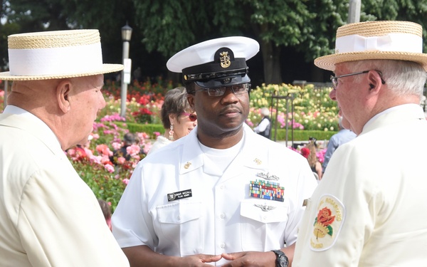 A U.S. Navy officer in white dress uniform is engaged in conversation with two civilians wearing cream-colored suits and straw boater hats at what appears to be an outdoor ceremonial event. The setting features a colorful rose garden in the background, suggesting this may be a Rose Festival or similar civic celebration. The Navy officer displays multiple service ribbons and medals on his uniform.