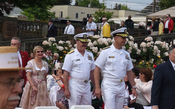 An outdoor ceremonial event featuring military officers in white dress uniforms alongside civilians in formal attire, set among blooming rose gardens. The scene appears to be a formal procession or reception with rows of white folding chairs arranged on a red carpet. The setting suggests a civic or commemorative ceremony, possibly a rose festival or garden dedication.