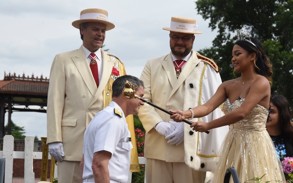A formal ceremonial knighting or investiture is taking place outdoors. A person in a white naval/military uniform kneels on a red cushioned stool while a young woman in a gold ballgown and tiara touches a ceremonial scepter to his shoulder. Two men wearing cream suits, red ties, medals, and straw boater hats labeled 'Royal Rosarians' stand as witnesses in the background.