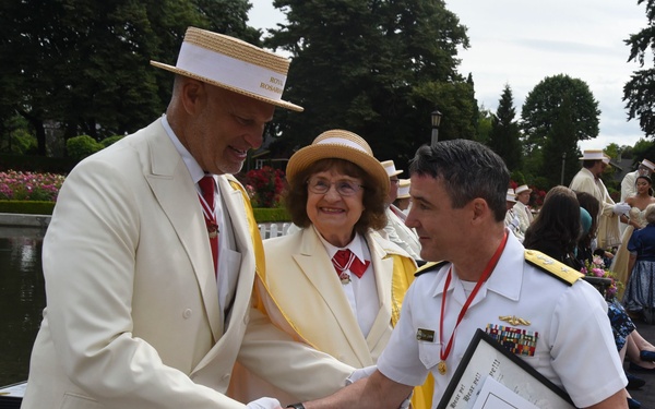 A formal outdoor ceremony appears to be taking place near a waterway, likely the Portland Rose Festival based on visible text on the straw hat reading 'Royal Rosarian.' Two individuals in cream/white suits with yellow sashes and straw boater hats are greeting a naval officer in white dress uniform who is holding framed certificates and a black hat.